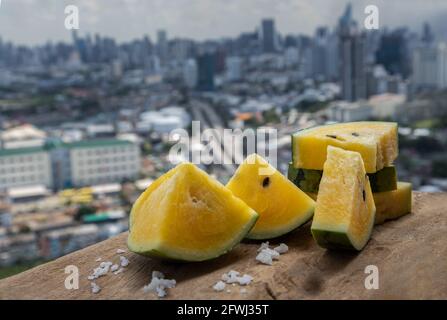 Fresh juicy yellow watermelon cut into triangles on old wooden table at balcony with city view. Tropical fruits, Select focus. Stock Photo