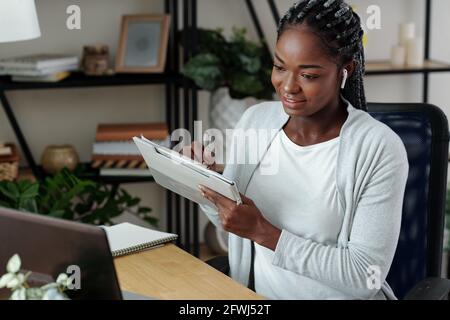 Smiling pretty woman watching webinar on laptop and taking notes, writing down important details Stock Photo