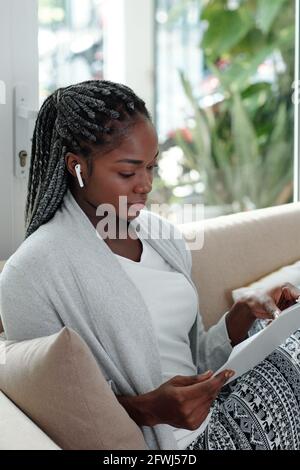 Pensive young woman in lounge clothes sitting on sofa, listening to music in earbuds and reading document Stock Photo