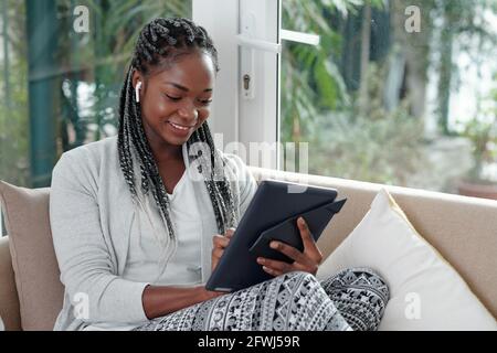 Happy young woman sitting on sofa at home and using application on tablet computer for communication with friends, family members and colleagues Stock Photo