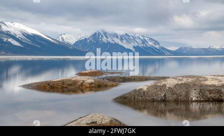 Scenic landscape in northern Canada during spring time with rocks, snow capped mountains reflection in calm lake water on cloudy afternoon. Stock Photo