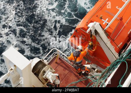 Aft hook on life boat fixed in  container vessel is checked by experienced crew member with orange jacket and white helmet. Stock Photo