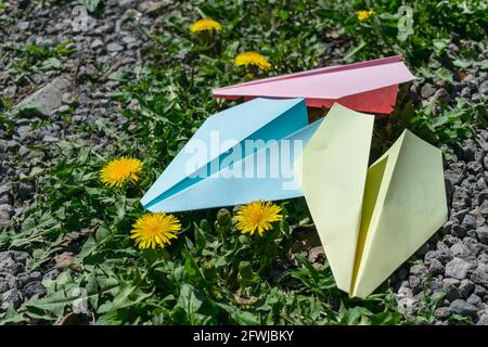Three multi-colored paper planes lie on the ground in the grass among yellow dandelion flowers. Children's creativity with origami Stock Photo