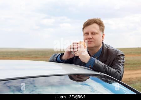 A man eats leaning on the roof of a car, against the blue sky. Stock Photo