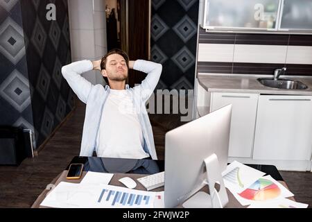 Happy young male worker resting on chair with folded hands behind head, closing eyes, enjoying break pause time during difficult workday in modern Stock Photo