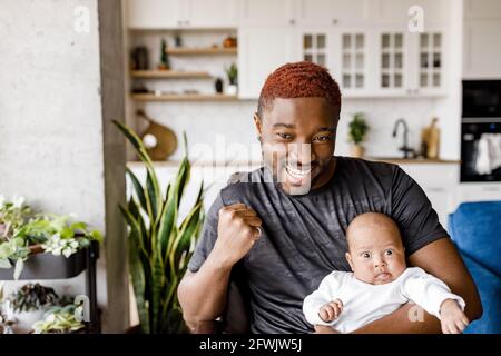 Happy young man sitting at home office room holding baby son, read message feel amazed by great online news. Happy male triumph reading pleasant good Stock Photo