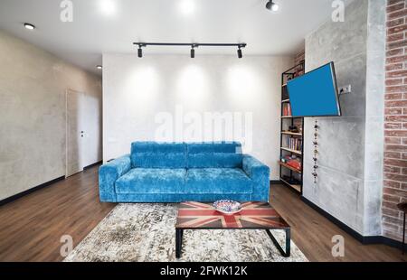 Stylish light living room interior with blue sofa, floor panels, interesting coffee table with image of british flag, TV set and shelves for accessories. Stock Photo