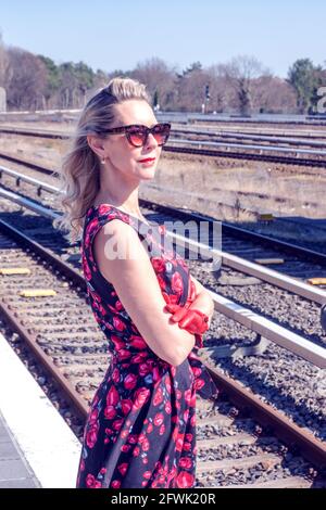 blonde woman in a red dress and sunglasses waiting at train station Stock Photo