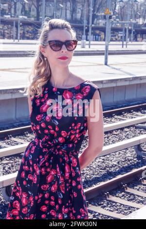 blonde woman in a red dress and sunglasses waiting at train station Stock Photo