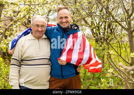 Handsome dad with his little cute sun are laying on green grassy lawn on American flag with American football ball in hand. Stock Photo