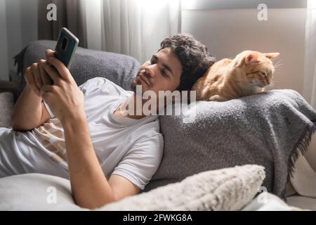 young man lays his head on the back of a brown tabby cat lying on the sofa, while using his cellphone Stock Photo