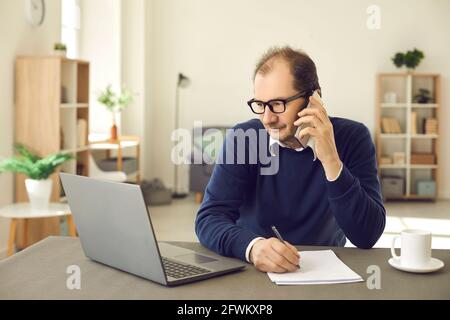 Mature multitasking man talking phone, looking at laptop making notes portrait Stock Photo