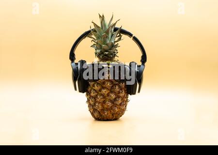 Funny pineapple in sunglasses wearing wireless headphones looking at camera over pastel yellow background. Party concept Stock Photo