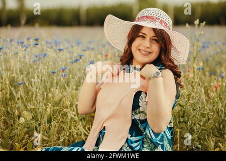 Close up of young smiling pregnant woman sitting on grass in field and holding her future baby clothing Stock Photo