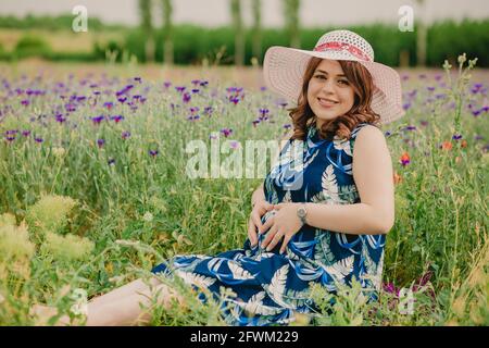 Smiling pregnant woman sitting on grass in field full of colorful flowers and touching her big belly Stock Photo