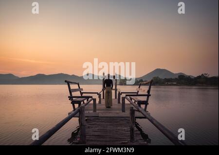 Young woman standing on wooden pier looking at sunrise on mountain at reservoir Stock Photo