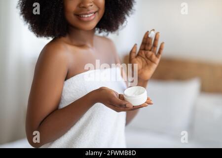 Attractive young black female in bath towel applying natural moisturizing cream on her face at home, closeup view Stock Photo