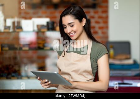 Young female owner of modern cafe in apron holding digital tablet and standing with shopwindow on background Stock Photo