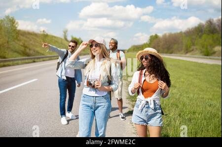 Group of millennial multiracial friends with backpacks walking along road, traveling together outdoors Stock Photo