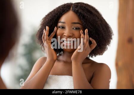 Lovely black lady standing in front of mirror, checking first wrinkles, touching her skin after applying cream at home Stock Photo