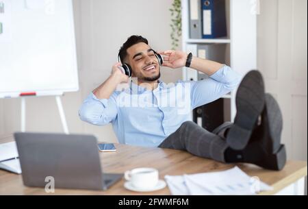 Taking break from work. Smiling Arab man listening to music in headphones, relaxing at workplace Stock Photo