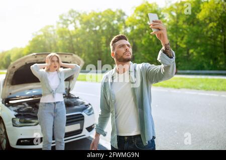 Sad young man standing on road next to broken car together with girlfriend, trying to call assistance. No connection Stock Photo