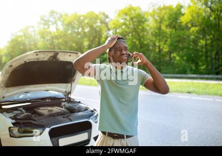 Unhappy black man standing near his broken car with open hood, calling breakdown road service on smartphone Stock Photo