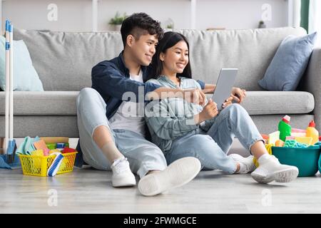 Loving asian couple with cleaning tools resting, using digital tablet Stock Photo