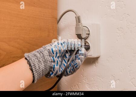 The hand of a male construction worker in protective gloves connects a plug to a double socket against the background of a wall. Stock Photo