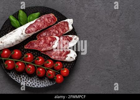 Sliced thin catalan fuet sausage and bunch of red cherry tomatoes on a black plate over dark rough surface. Traditional spanish dry-cured pork salami. Stock Photo