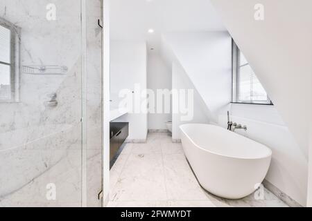 Interior of spacious bathroom in marble tiles with soaker tub near window in attic Stock Photo