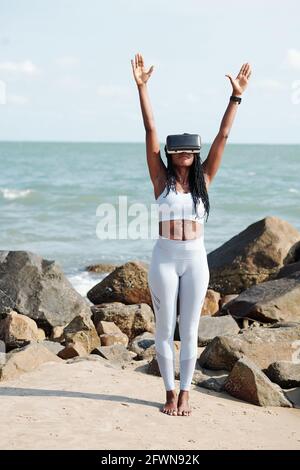 Fit young woman in virtual reality headset working out on beach on sunny day Stock Photo