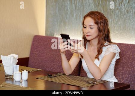 Pretty young woman sitting at restaurant table, texting friends or checking social media Stock Photo