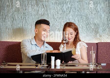 Joyful young couple sitting at restaurant table, reading menu and choosing food for dinner Stock Photo