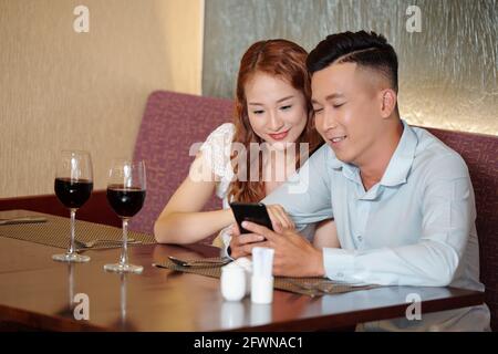 Happy young couple sitting at cafe table, drinking wine and reading interesting post on social media or watching new video of famous blogger Stock Photo