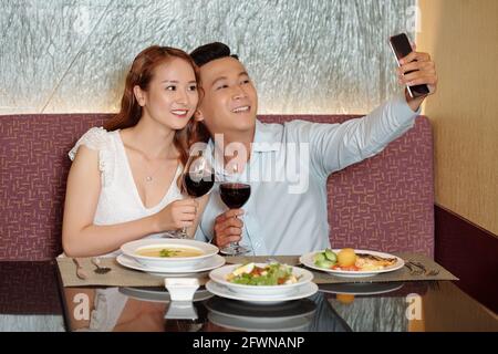 Smiling young couple taking selfie with wine glasses at table in fancy restaurant when having romantic dinner Stock Photo