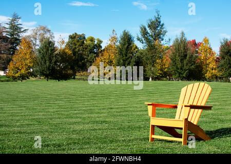 Traditional curveback sunset plastic outdoor patio adirondack chair with contoured backs and seats on green grass of lawn surrounded by autumn trees