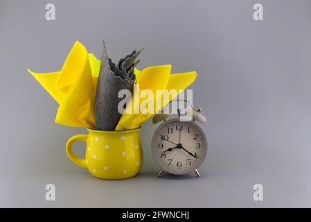 Yellow and gray napkins placed in the yellow cup next to the gray vintage alarm clock on gray background with space for text Stock Photo