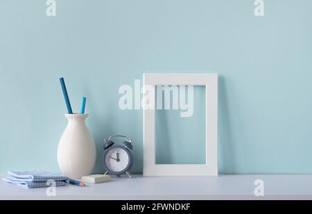 Interior with blue tone. Workplace of designer. White wooden blank frame on a desk, alarm watch, vase with pencils. Mockup with place for text. Stock Photo