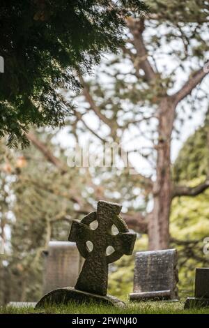Dark moody graveyard scene with leaning cross headstone in old Victorian churchyard.  Dark and eerie light with sinister feeling. Stock Photo