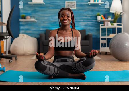 Fit black woman practicing yoga sitting with legs crossed in sportive top and leggins on lotus pose. Practicing calm mind harmony for no stress life in home living room. Stock Photo