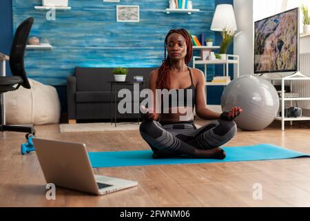 Afro american woman in sportwear sitting practicing lotus position doing yoga sitting on yoga mat dressed in sportwear following online training. Workout and healthy lifestyle. Stock Photo