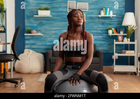 Tired fit black woman after working out in home living room, resting using stability swiss ball, wearing sportive outfit. Training indoors for muscular growth. Stock Photo