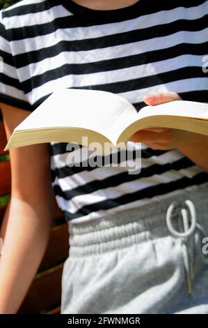 Person sitting on the bench, holding a white book and reading outdoors. Leisure time in the garden. Stock Photo