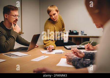 Female executive pointing at paper and discussing project with creative team in meeting. Team sitting around table and discussing creative ideas in me Stock Photo