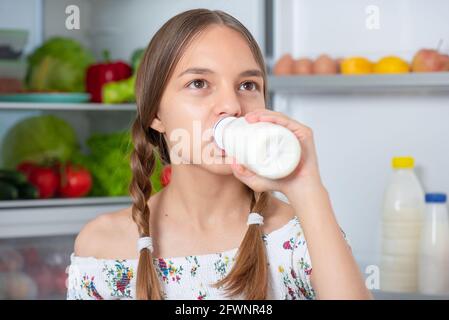 Beautiful young teen girl holding bottle of milk and drinks while standing near open fridge in kitchen at home Stock Photo