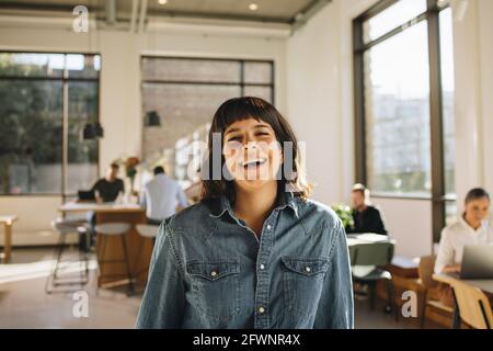 Portrait of happy freelancer standing in co-working space with people working in background. Happy businesswoman laughing in office. Stock Photo