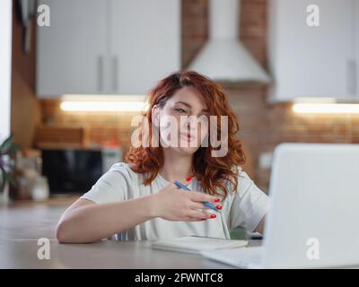 A beautiful young redheaded woman sits in the kitchen at the table with her laptop. Stock Photo