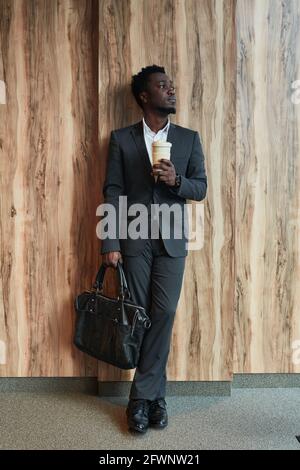 Vertical full length portrait of successful African-American businessman looking away while standing by wood background Stock Photo