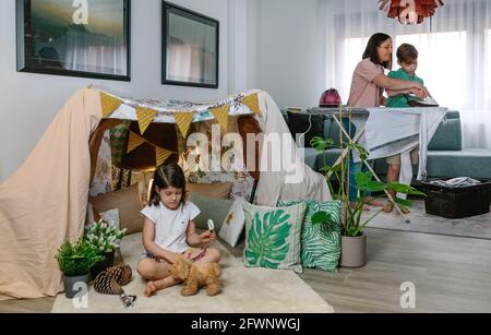 Mother teaching her teenage son to iron while her daughter play camping at home Stock Photo